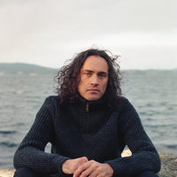 Man with curly hair wearing a dark sweater sitting on a rock by a body of water.