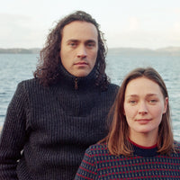 Man with long hair wearing a dark sweater sitting by a body of water.