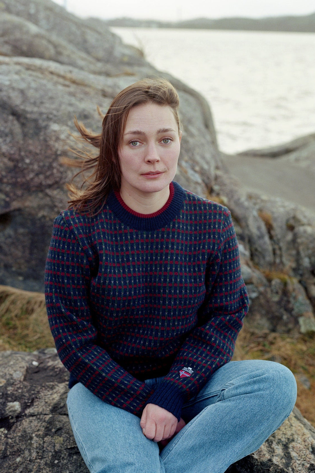 Person sitting on rocks by a lake wearing a patterned sweater and jeans.