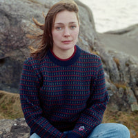 Person sitting on rocks by a lake wearing a patterned sweater and jeans.
