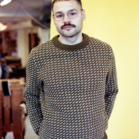 Man wearing a patterned sweater and glasses standing against a yellow wall.
