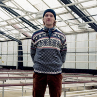 Person wearing a patterned sweater and brown pants standing inside a greenhouse.