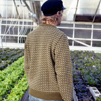 Person wearing a patterned sweater and cap in a greenhouse with plants.