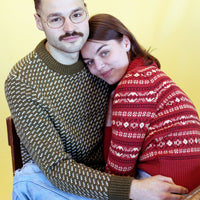 Two people sitting close together wearing patterned sweaters against a yellow wall.