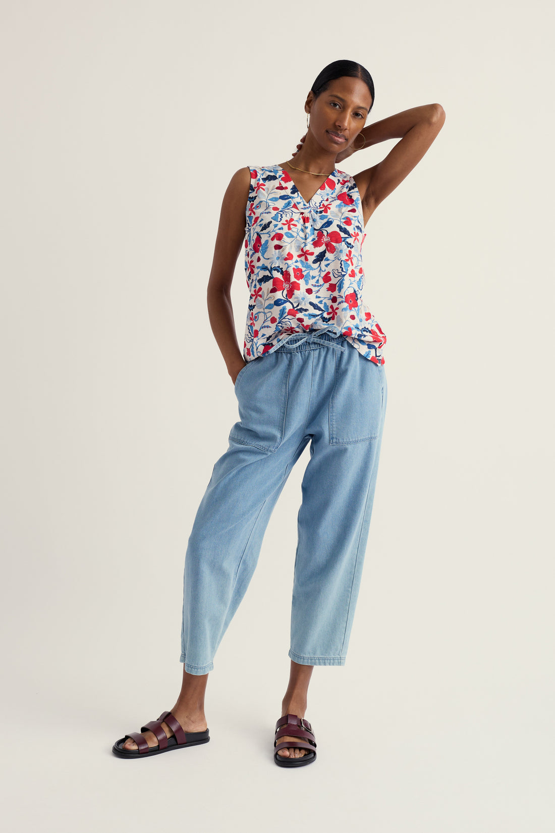 Woman wearing a floral sleeveless top and light blue pants on a white background