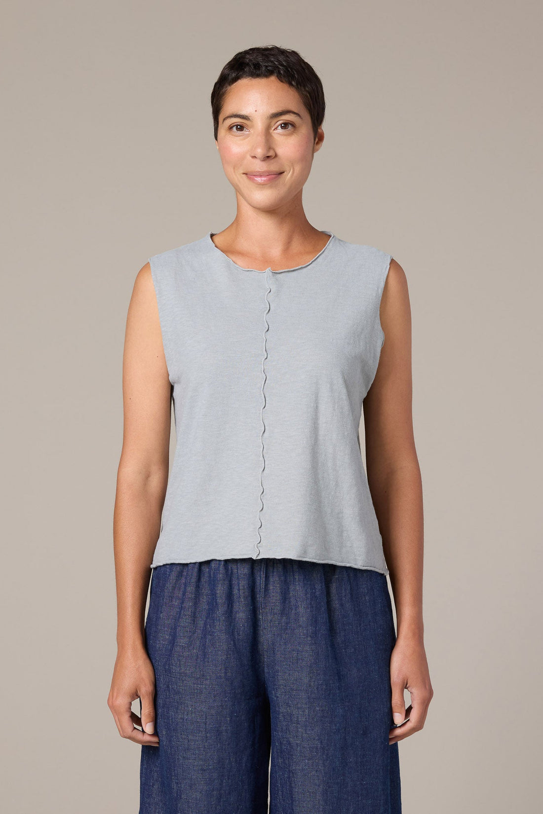 Woman wearing a light blue sleeveless top and dark blue pants against a beige background