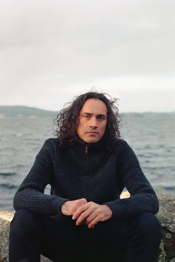 Man with curly hair wearing a dark sweater sitting on a rock by a body of water.