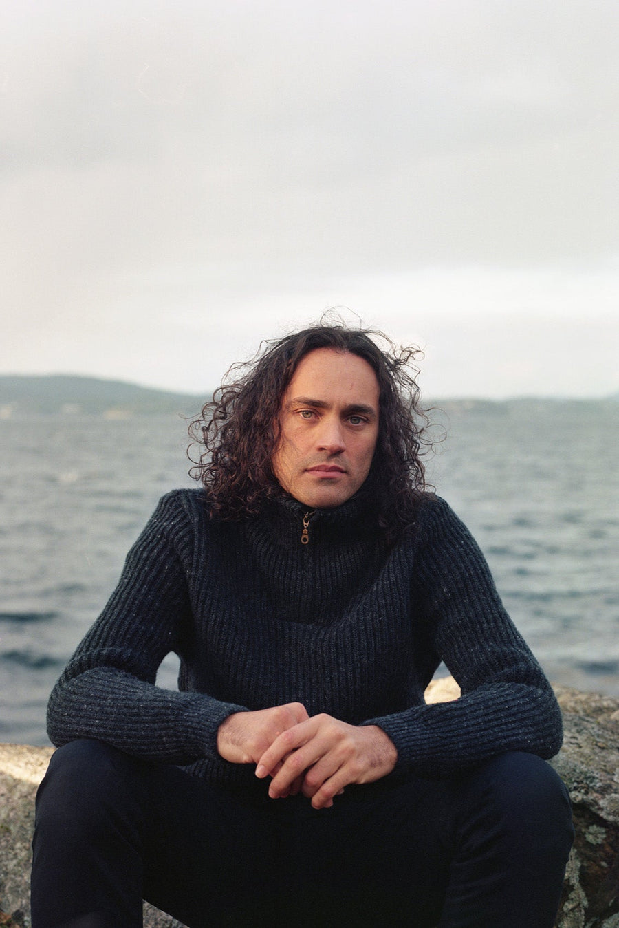 Man with curly hair wearing a dark sweater sitting on a rock by a body of water.