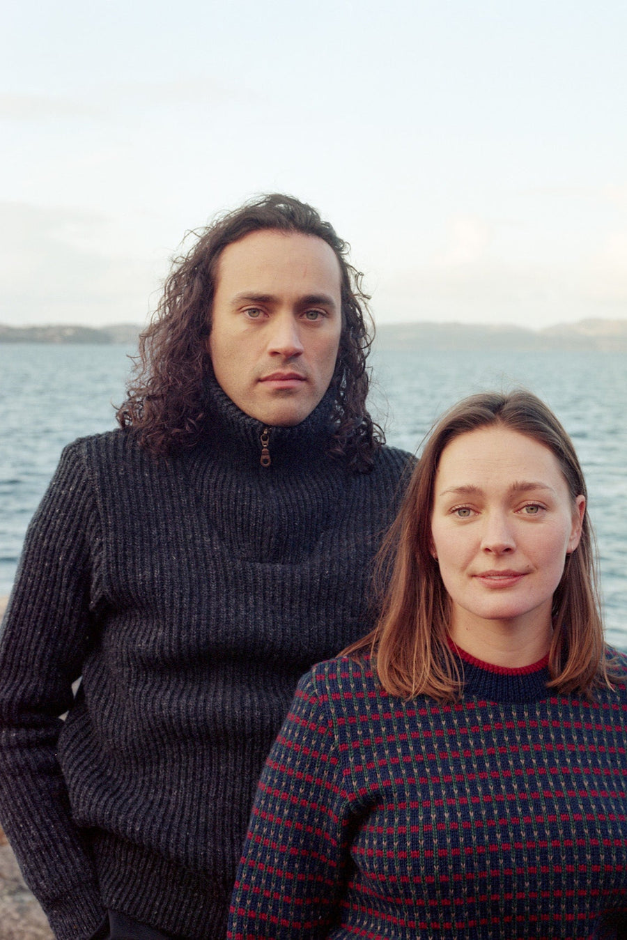Man with long hair wearing a dark sweater sitting by a body of water.