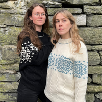 Two women standing against a stone wall, one wearing a black sweater with white patterns and the other in a white sweater with blue patterns.