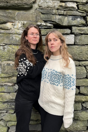 Two women standing against a stone wall, one wearing a black sweater with white patterns and the other in a white sweater with blue patterns.