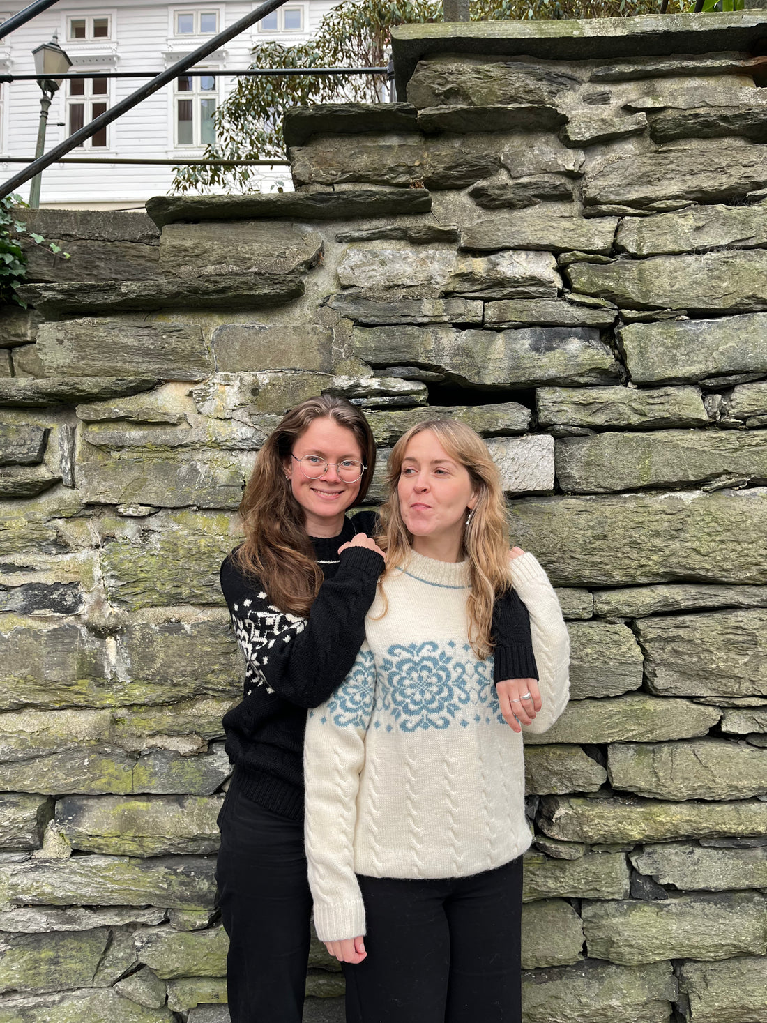 Two women standing against a stone wall with a house in the background