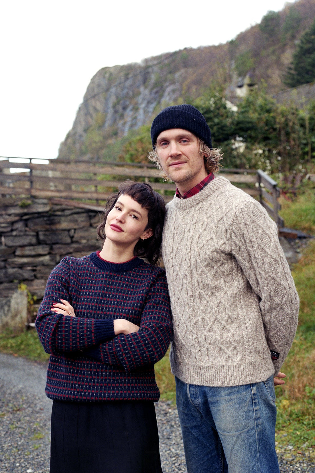 Two people standing outdoors with a scenic background of mountains and trees.