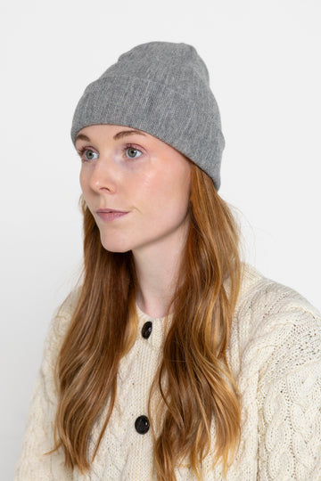 A person wearing a gray beanie hat and a white knitted cardigan against a neutral background.