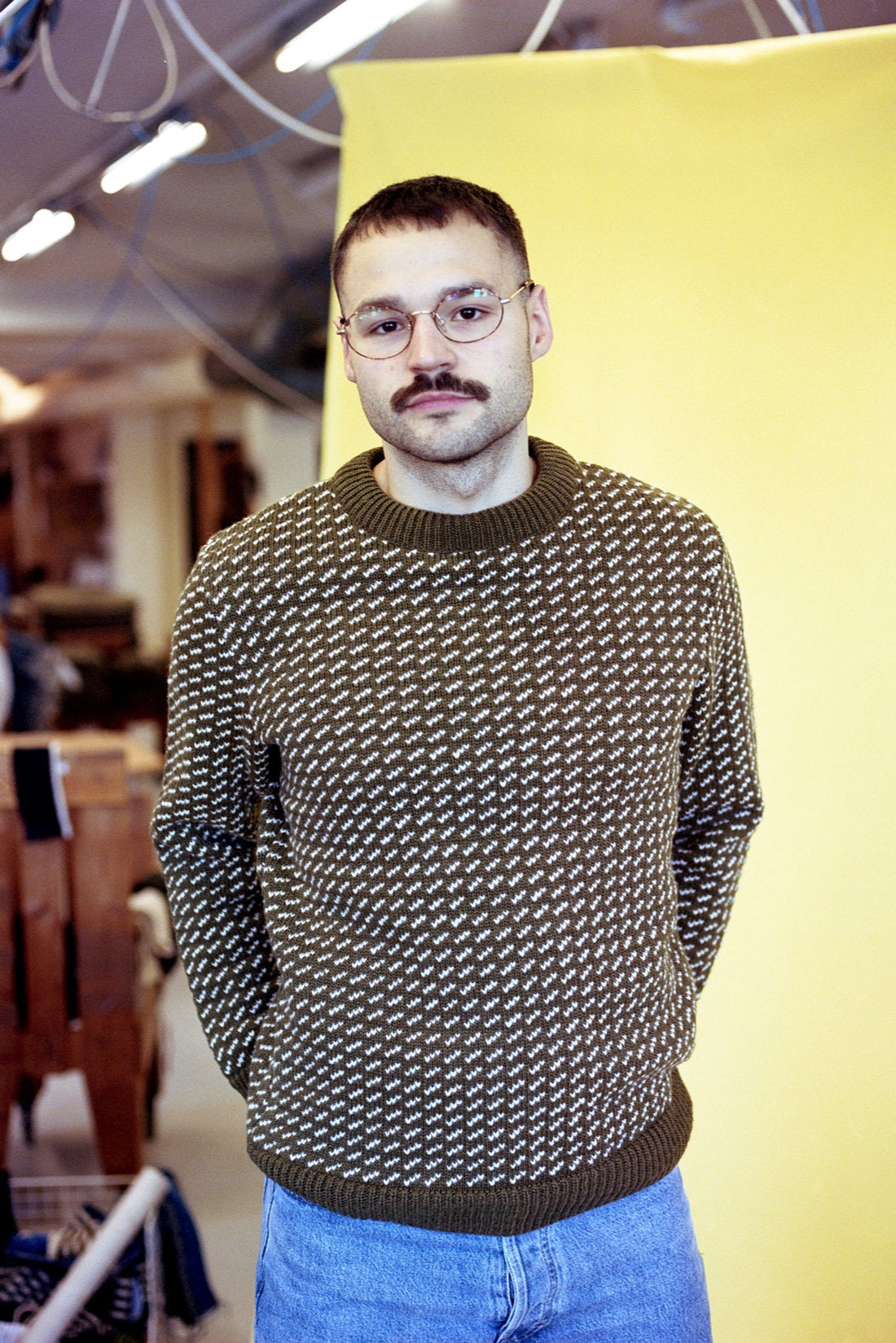 Man wearing a patterned sweater and glasses standing against a yellow wall.