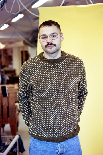 Man wearing a patterned sweater and glasses standing against a yellow wall.