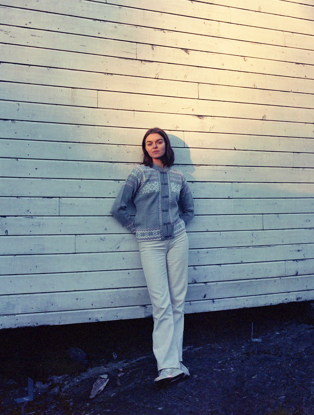 Model wears the Voss cardigan in Grey and leans against a wooden wall.