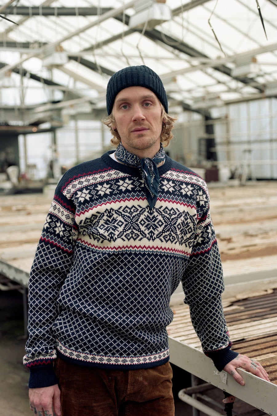 Man wearing a patterned sweater and beanie standing in a greenhouse.