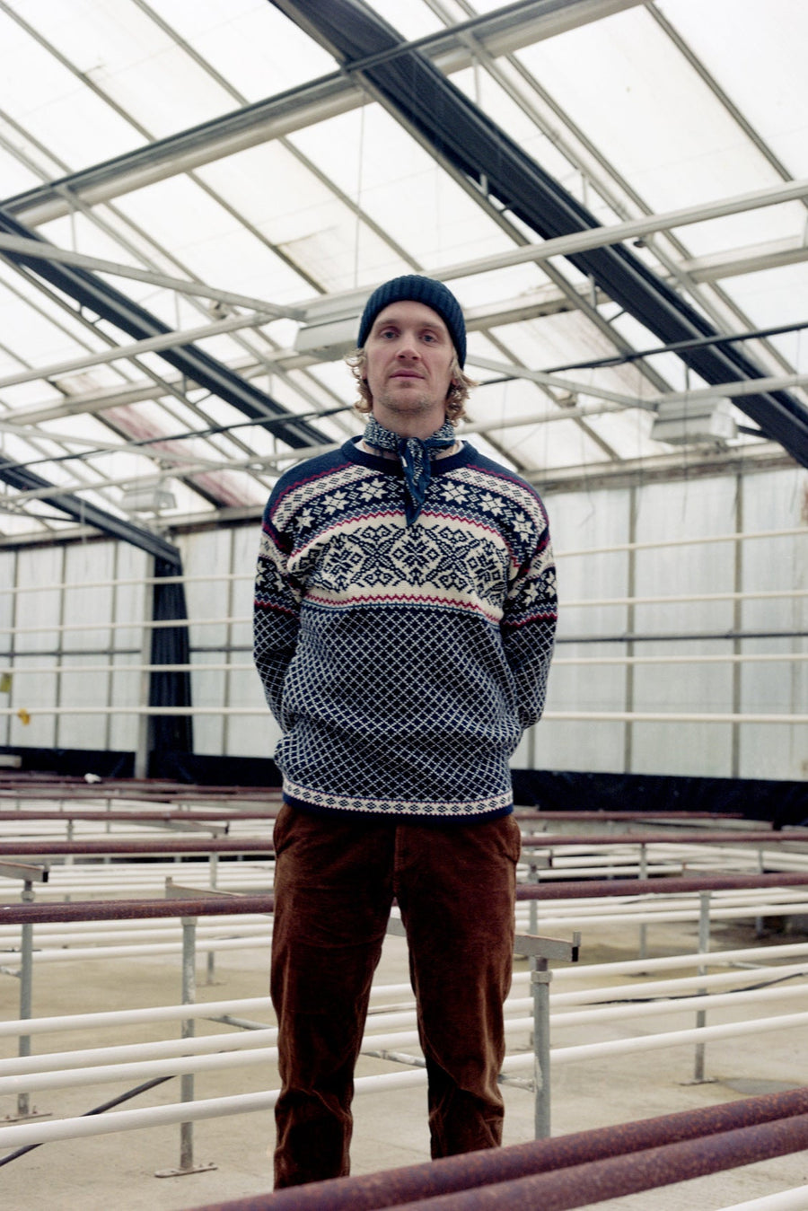 Person wearing a patterned sweater and brown pants standing inside a greenhouse.