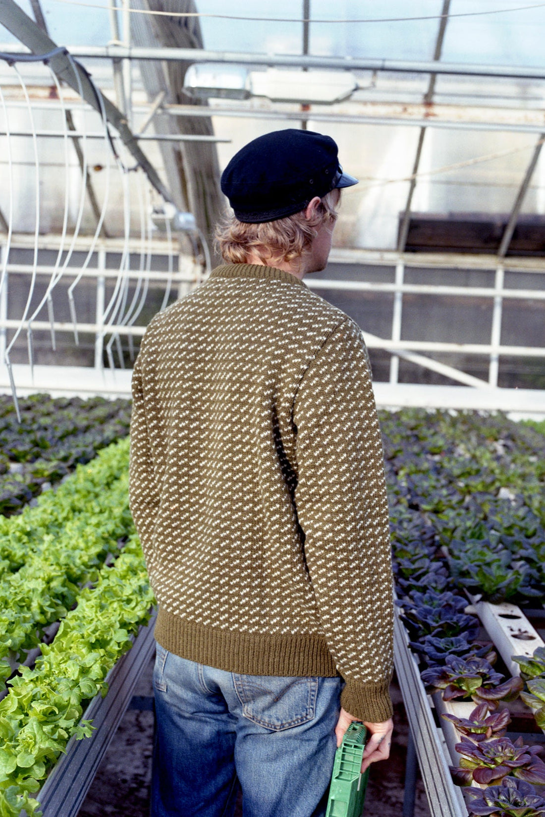 Person wearing a patterned sweater and cap in a greenhouse with plants.