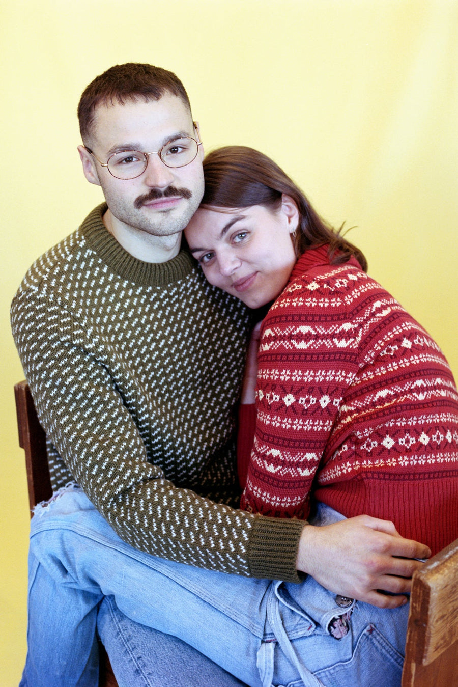 Two people sitting close together wearing patterned sweaters against a yellow wall.